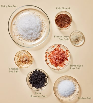 Assorted gourmet salts in bowls: pink, black, grey, and flaky varieties on a beige background.