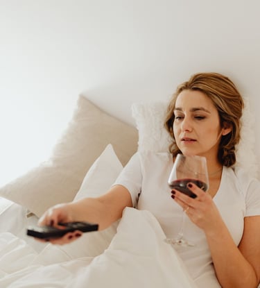 A woman relaxes in bed holding a glass of red wine and using a TV remote.