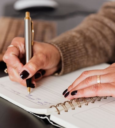 A person writing in a spiral notebook with a silver pen at a desk.