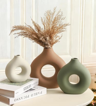 Modern donut vases in ceramic matte finishes with dried pampas grass on a white side table.