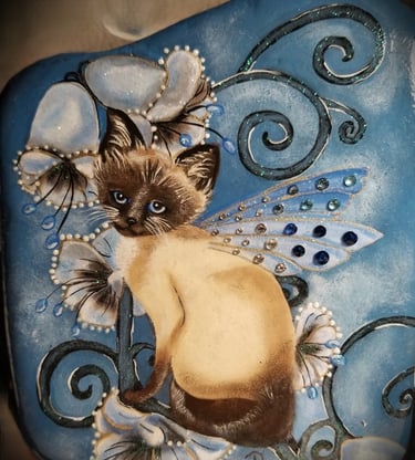 A rock painting of a Siamese Fairy Kitty on blue bell flowers.