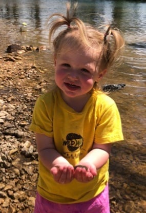 A toddler girl with pigtails holding fish while standing on a pebble riverbank during a summer day.