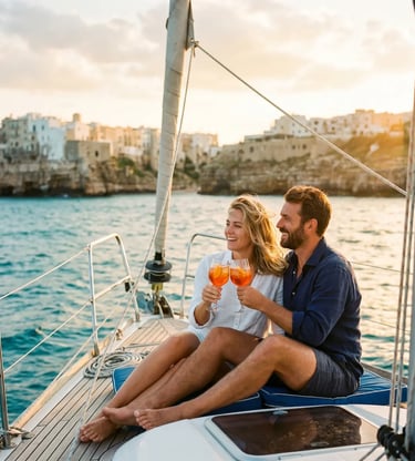 Couple toasting with Aperol Spritz on sailboat, Polignano a Mare cliffs in background