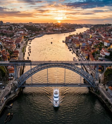 Aerial view of a yacht passing under Dom Luís Bridge on the Douro River at sunset.