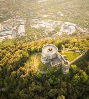 Drone shot of medieval castle ruins surrounded by green forest and urban sprawl