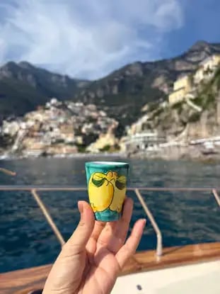 Hand holding traditional lemon ceramic cup on boat with Positano village and mountains in background