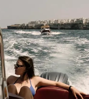 Woman in bikini lounging on speedboat cruising past whitewashed Polignano a Mare cliffs