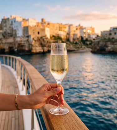 Hand holding chilled Apulian prosecco glass on yacht deck, Polignano a Mare cliffs at golden hour