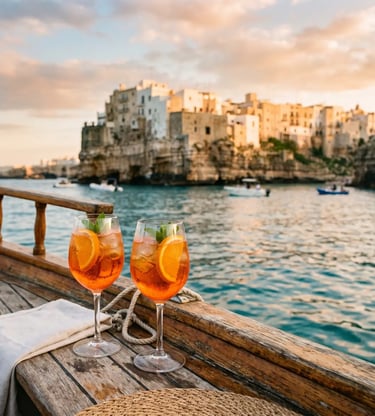 Two Aperol spritz glasses on boat deck with Polignano a Mare at sunset