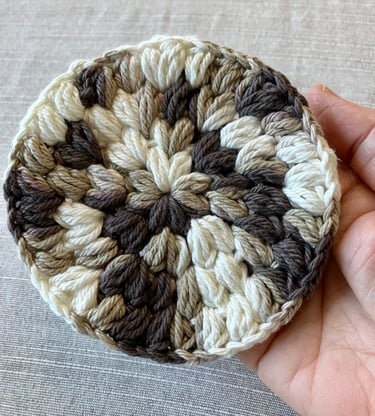 A hand holding a brown, tan, and cream colored crocheted drink coaster.
