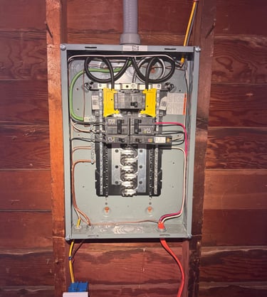 Open electrical service panel showing breakers and wiring mounted on a wood wall.