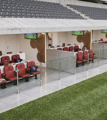 Luxury stadium VIP boxes with red leather seats, private dining tables, and TVs overlooking a football pitch.