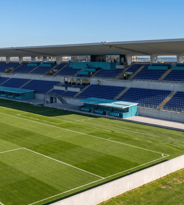 Empty outdoor football stadium with blue seats and green grass field under a clear blue sky.