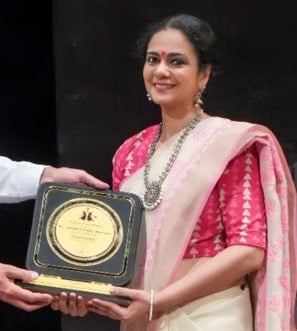 a woman in a white and pink saree receiving an award. She is Ms. Anushree Ladia, Bharatnatyam Expert