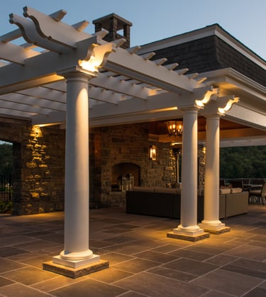 Luxury outdoor patio with stone fireplace, lit white pergola, and bluestone pavers at dusk.