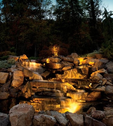 Illuminated backyard rock waterfall feature with warm LED landscape lighting at night.