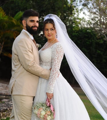 Uma noiva em um vestido de renda e véu longo posando ao ar livre com seu noivo de terno bege.