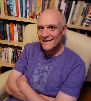 Gregory Spatz sits in an armchair in front of a bookshelf. He wears a purple shirt and smiles.