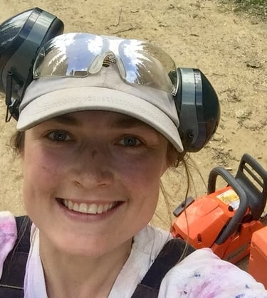 Keely smiles wide, wearing a beige ballcap and headphones. Behind her is an orange chainsaw.