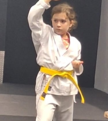 Young girl in a white karate gi with a yellow belt practicing a high block technique in a martial arts dojo.