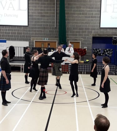 Dancers, dressed mostly in black, dancing a strathspey circle. Judges in the background on a stage.