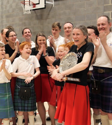 Laughing woman clutching a large trophy. Her team surrounds her, smiling and clapping.