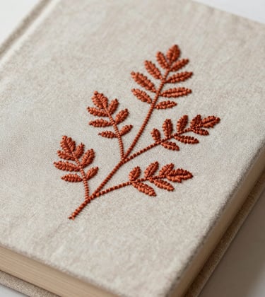 A close-up photograph of a vintage linen book cover featuring delicate, hand-stitched botanical embroidery in terracotta thread. Calm, artistic composition for a luxury interior brand.