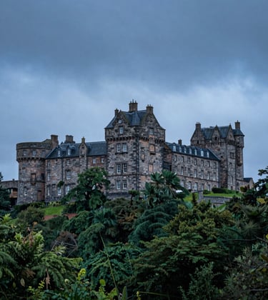 A wide-angle photograph of a historic stone castle in Scotland during a moody blue hour, with deep forest green trees and soft cloud white mist.