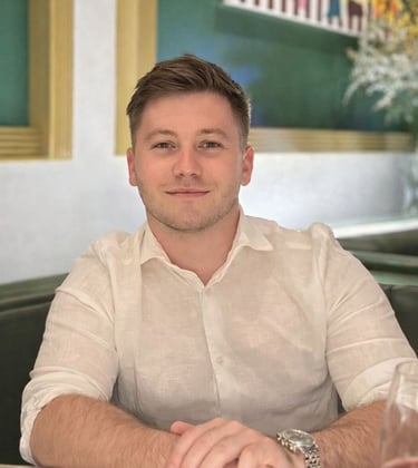 Harrison, smiling, wearing a white linen shirt and silver watch sitting at a restaurant table.
