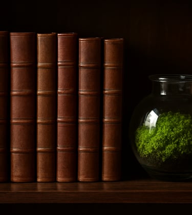 A collection of scholarly journals and textbooks bound in Wood Brown leather, arranged neatly on a dark wooden shelf next to a small glass terrarium containing lush green moss.