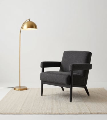 A minimalist living area with a single charcoal armchair, a cream textured rug, and a soft gold floor lamp in a clean white space. International / High-end residential photography.