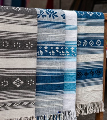 Close-up photography of an artisan market in Oaxaca, Mexico. Traditional textiles with charcoal and soft blue patterns are draped over wooden stalls under natural, diffused lighting.