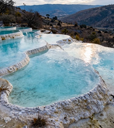 A high-angle shot of the turquoise mineral pools perched on the edge of the cliff at Hierve el Agua, Oaxaca, Mexico. The water is a crystal clear light blue, reflecting the afternoon sun. The surrounding Mexican landscape of dry vegetation and distant blue mountains creates a contemplative atmosphere.