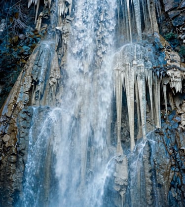 Photography of the petrified waterfall's vertical face at Hierve el Agua. The mineral columns create a dramatic, stalactite-like texture in shades of off-white and soft blue against the mountain shadow.