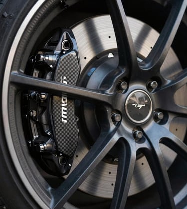 A close-up studio photograph of a high-performance carbon-ceramic brake and a multi-spoke alloy wheel. The metal surfaces reflect a soft, cool off-white glow, with deep shadows emphasizing the mechanical precision and obsidian black finish.
