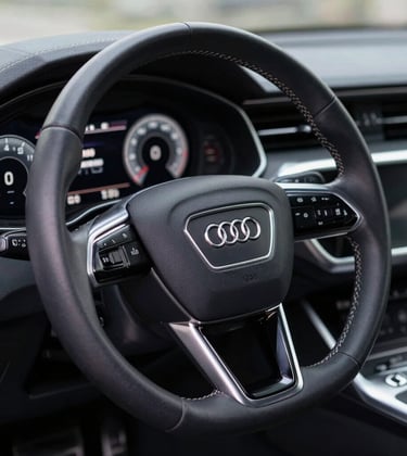 A sharp, high-detail macro photograph of an Audi's digital dashboard and leather-wrapped steering wheel. The interface glows with muted slate blue light, set against a dark, sophisticated interior with cool off-white stitching details.