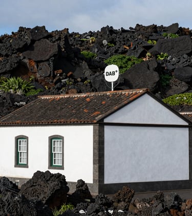 A traditional white-walled Atlantic island building with a discrete modern DAB+ antenna on its roof, surrounded by volcanic rocks and greenery in the Azores, European Atlantic / Insular.