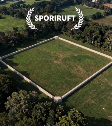 Clean, professional drone shot focusing on the boundary markings of the 1-acre site. The land is flat and cleared, framed by soft off-white concrete markers against the rich green and navy tones of the surrounding landscape.