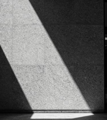 A minimalist, high-contrast black and white photograph of a single shaft of light illuminating a granite texture on a wall in a North American / International Business lobby. Sharp shadows, intellectual precision.