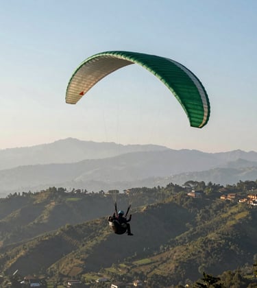 A wide-angle shot of a paraglider in flight over the lush hills of Godawari, Lalitpur. In the background, the distant Himalayan range is visible under a clear sky. The paraglider wing has accents of muted forest green.