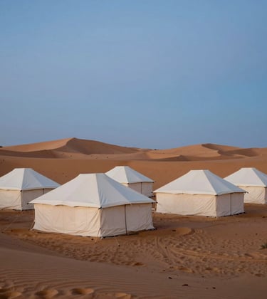 A luxury desert camp setting in a North African / Moroccan landscape during the blue hour. High-end creamy off-white canvas tents are arranged neatly against soft warm sand dunes. Minimalist composition with a focus on silence and space.