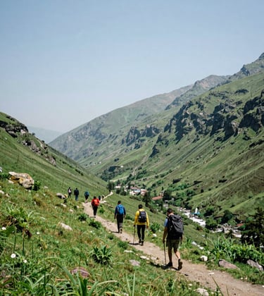 A bright outdoor photograph of a lush green mountain valley in South Asia / India, with hikers on a trail under a pale blue sky, crisp natural light.
