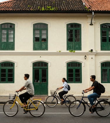 A premium photograph of the Fatahillah Square in Jakarta's Batavia Old Town. The scene features colonial architecture with ivory walls and mossy leaf green window frames. Southeast Asian / Indonesian travelers are exploring the plaza on colorful vintage bicycles.