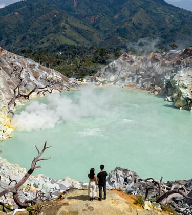 A stunning landscape photo of Kawah Putih in Bandung. The volcanic crater lake is a pale seafoam green, contrasted with white soil and skeletal trees. Wisps of steam rise against a deep forest green mountain backdrop. A Southeast Asian / Indonesian couple is visible taking in the view.