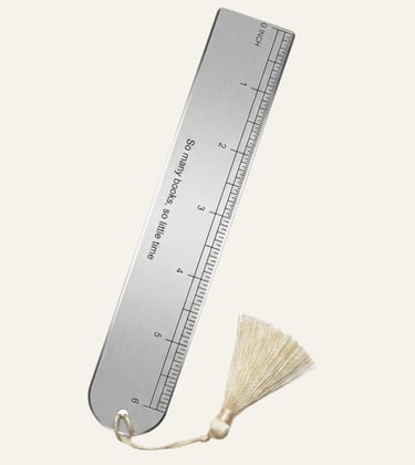 A brushed metal ruler bookmark with an engraved quote and a cream silk tassel on a plain background.