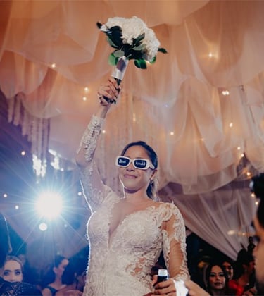 Novia levantando ramo de boda en la mano derecha con lentes de sol con las iniciales de ella y novio