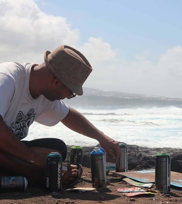 Artist in a fedora hat preparing spray paint cans for a mural on a rocky coastline.