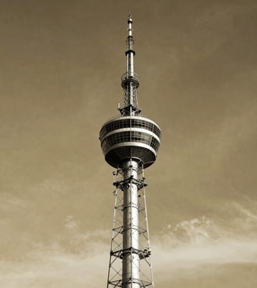 A black and white style professional photograph with warm gold toning, showing a classic broadcast tower standing tall against a dramatic sky in Romania. Graphic, elegant composition, capturing the historical weight and early days of wireless transmission. Sophisticated and scholarly aesthetic.