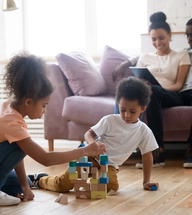 a girl and boy playing with blocks on the floor with parents planning in the background