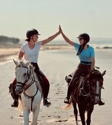 2 personnes souriantes montant à cheval sur la plage en se tapant dans la main 